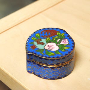 May include: A small, blue, oval-shaped trinket box with a floral design on the lid. The box features a detailed pattern with gold trim and a vibrant color palette of blue, red, green, and pink. The box is sitting on a light-colored wooden surface.