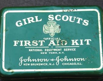 Könnte beinhalten: Eine rechteckige grüne Metalldose für ein offizielles Erste-Hilfe-Set der Girl Scouts. Der Deckel trägt den weißen Schriftzug Girl Scouts Official First Aid Kit, National Equipment Service, New York, N.Y. sowie Johnson & Johnson, New Brunswick, N.J., Chicago, Ill.