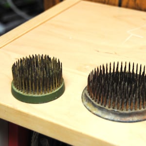 Op de afbeelding: Twee bloemenspinnen, ook wel kenzans genoemd, liggen op een licht houten oppervlak. De ronde gereedschappen hebben dicht op elkaar geplaatste, scherpe metalen pinnen. Eén heeft een groene basis, de andere een zilveren. Ze worden gebruikt in bloemstukken.
