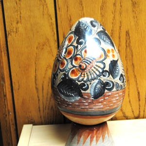 May include: A decorative, egg-shaped ceramic vase with a floral design in orange, black, and cream colors. The vase sits on a pedestal base with a gray and orange pattern. The background is a wooden wall.