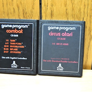 May include: Two black Atari game cartridges with white text. The left cartridge is for the game "Combat" and has the game code CX-2601. The right cartridge is for the game "Circus Atari" and has the game code CX-2630.