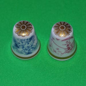 Puede incluir: Dos dedal de porcelana blanca con ribetes dorados y diseños florales. Un dedal tiene un diseño floral azul y el otro tiene un diseño floral rosa.