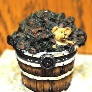 May include: A decorative barrel-shaped trinket box with a teddy bear peeking out from a bed of leaves. The box has a wooden appearance with white stripes and a paw print clasp. The leaves are green and red.