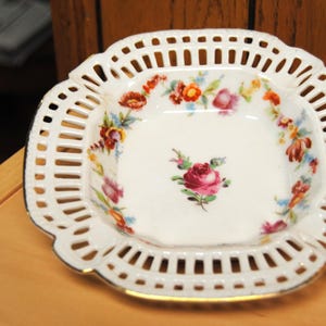 May include: White porcelain dish with a pierced, scalloped edge and floral design. The square dish features a central rose and colorful flowers around the rim. The rim has a gold trim.