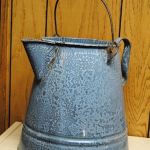 May include: Vintage blue speckled enamelware pitcher with a spout and a wire handle with a black wooden grip. The pitcher has a rounded base and a slightly flared top. The handle is attached to the sides of the pitcher.
