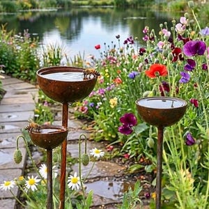 May include: Three weathered metal bird baths of different heights are positioned in a garden, each holding water. Bees are visible on the rims. The garden is filled with colourful flowers, with a pond visible in the background.