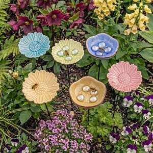 Pode incluir: Seis estacas de jardim em cerâmica com formato de flor em cores pastel, incluindo azul claro, amarelo e rosa, montadas em hastes de metal preto. Vários dos pratos de cerâmica contêm pedras de vidro transparentes e pequenas abelhas.