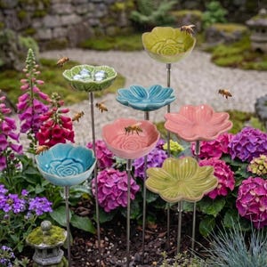 Könnte beinhalten: Eine Sammlung von sieben blumenförmigen Gartensteckern aus Keramik in Hellgrün, Blau und Rosa, die jeweils auf einem dünnen Metallstab montiert sind. Die Schalen haben geprägte Blütenblattmuster, einige enthalten kleine Glaskugeln und Bienen.