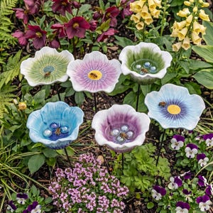 May include: A selection of ceramic flower-shaped bird baths in shades of blue, green, and purple, with bees and glass beads. The bird baths are on black metal stems, placed in a garden setting with flowers and foliage.