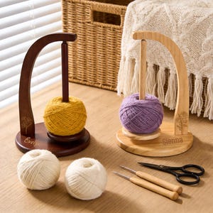 May include: Two wooden yarn holders with yarn balls in yellow and purple. The holders have a curved arm and a central dowel. Two white yarn balls, two crochet hooks, and a pair of scissors are also on the wooden surface.