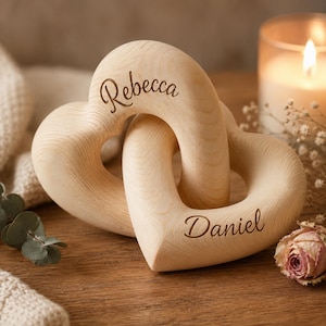 Könnte beinhalten: Zwei ineinandergreifende Holzherzformen, graviert mit den Namen "Rebecca" und "Daniel". Die Herzen sind aus hellfarbenem Holz mit glatter Oberfläche. Eine brennende Kerze und Trockenblumen befinden sich im Hintergrund.