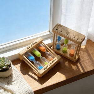 Op de afbeelding: Twee houten zandlopers met blauw, groen en oranje zand. Eén zandloper heeft de tekst "Olivia & Noah" en "Forever together". De andere zandloper heeft de tekst "1 MIN", "3 MIN" en "5 MIN".