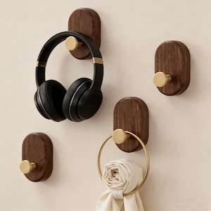 May include: Four wooden wall hooks with brass knobs. One hook holds black headphones, and another holds a gold towel ring with a cream towel. The oval-shaped hooks are mounted on a light-coloured wall.