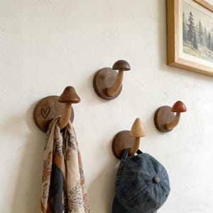 May include: A collection of wooden mushroom-shaped wall hooks in shades of brown. One hook holds a patterned scarf, another a blue baseball cap. One hook has a heart carved into the wooden base. The hooks are mounted on a white wall.