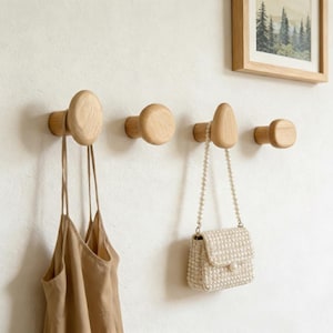May include: Four wooden wall hooks of different shapes are fixed to a white wall. A beige dress hangs from the first hook, and a small, white beaded purse hangs from the third hook. A framed picture of a forest scene is above the hooks.