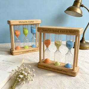 Può includere: Set di clessidre in legno con tre tubi di vetro riempiti di sabbia arancione, verde e blu. La cornice in legno riporta la scritta "Sand in hand, love in heart" e "Time gentle, love eternal". Un oggetto decorativo.
