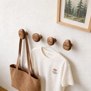 May include: Three wooden wall hooks, each engraved with the name "Olivia". A brown tote bag and a white t-shirt with a mushroom design are hanging from the hooks. A framed picture of trees is visible on the wall.