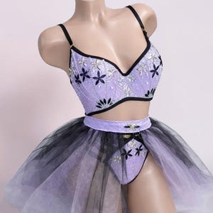 May include: A lavender lingerie set with black detailing. The bra and briefs feature floral embroidery and silver embellishments. The set includes a layered tulle skirt that fades from black to lavender.