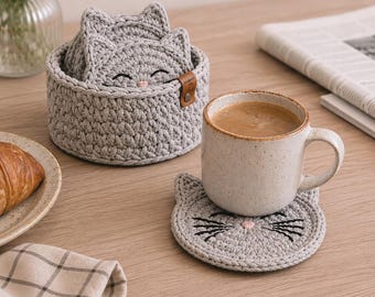 May include: A set of gray crochet cat-shaped coasters with embroidered details, including whiskers and a pink nose. The coasters are displayed with a coffee mug and a basket holding additional coasters. A croissant and flowers are also present.