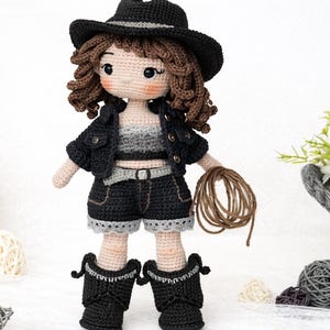 May include: A handmade crochet doll wearing a black hat, jacket, shorts, and boots. The doll has brown curly hair and is holding a brown rope. The doll's outfit includes a gray and white top and a belt. The doll is approximately 12 inches tall.