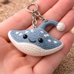 May include: A handmade blue and white crochet keychain in the shape of a smiling stingray. The keychain has a silver chain and ring. The stingray has white polka dots and black eyes. The background is a sandy beach.