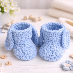 May include: A pair of light blue crochet baby booties with bunny ear details. The handmade booties are displayed on a white surface with decorative elements, including lace, wooden heart-shaped buttons, and floral accents. A ball of blue yarn is in the background.