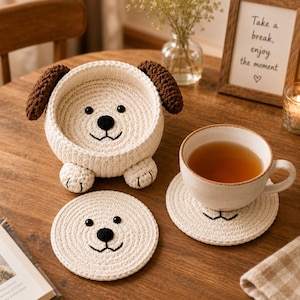 May include: A cream-colored crochet dog-shaped bowl with brown ears and two matching circular coasters. The bowl features a black embroidered nose, eyes, and mouth, with two small front paws at the base. One coaster is plain, and the other supports a ceramic mug.