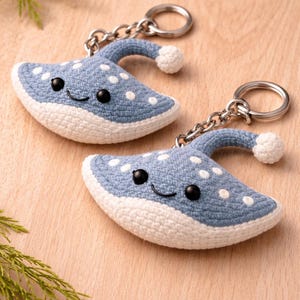 May include: Two handmade blue and white crochet stingray keychains with smiling faces and white polka dots. Each keychain has a silver chain and ring. The keychains are on a light brown wooden surface.
