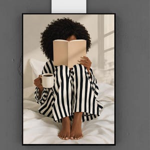 May include: A framed wall art print depicting a person with dark curly hair sitting on white bedding, wearing black and white striped pajamas. The person holds an open beige book in front of their face and a white mug in one hand, creating a scene.