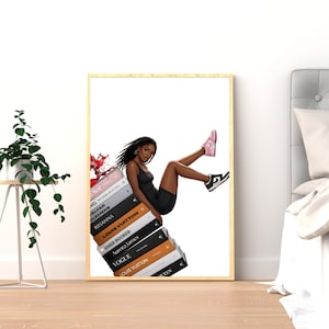May include: A framed art print featuring a stylish illustration of a person in a black outfit and sneakers, leaning on a stack of fashion books. The books include titles like Dior, Louis Vuitton, and Vogue. The print has a gold frame.