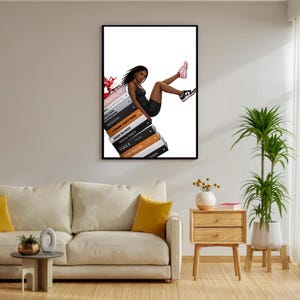 May include: A framed art print featuring a woman in a black dress and sneakers, seemingly perched atop a stack of fashion-related books. The books include titles like "Vogue" and "Louis Vuitton." The artwork has a white background and is displayed in a living room setting.