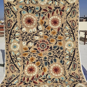 Op de afbeelding: Beige textiel met ingewikkelde bloemenborduurwerk. Het ontwerp heeft een centraal medaillon en herhaalde bloempatronen in blauw, rood, geel en wit. De rand is donkerbruin met een witte rand.