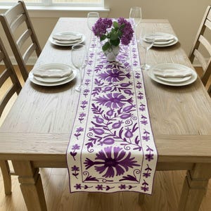 Może przedstawiać: Drewniany stół jadalny nakryty do posiłku, z białym i fioletowym haftowanym bieżnikiem w kwiaty. Stół jest nakryty białymi talerzami, kieliszkami do wina i wazonem z fioletowymi kwiatami. Krzesła są drewniane.