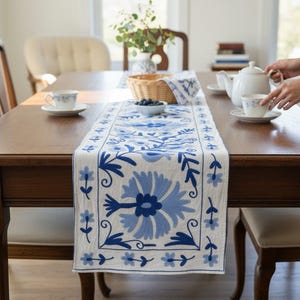 Puede incluir: Una mesa de comedor de madera con un camino de mesa bordado con flores azules y blancas. La mesa está puesta con tazas de té blancas y una tetera. La escena está iluminada por luz natural, creando un ambiente cálido y acogedor.