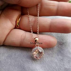 Puede incluir: Collar de oro rosa con un colgante redondo de color melocotón rodeado de pequeñas piedras transparentes. El colgante cuelga de una delicada cadena. El texto "Silver Dew" es visible en la imagen.