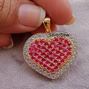 Puede incluir: Colgante en forma de corazón con una fianza dorada. El colgante presenta un centro de pequeñas piedras preciosas rojas rodeado por un borde de piedras transparentes. El borde exterior es dorado y el texto "SilverDew" es visible.