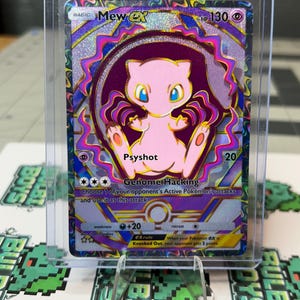 May include: A holographic Mew EX Pokémon trading card, encased in a protective sleeve. The card features a pink Mew character with blue eyes, set against a purple and silver background. Text includes "Psyshot" and "Genome Hacking".