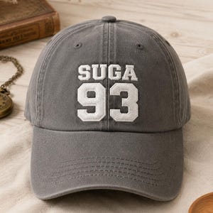 May include: Gray baseball cap with the text "SUGA" above the number "93" in white block letters. The cap has a curved brim and a fabric strap closure. The hat is made of a soft, durable material.