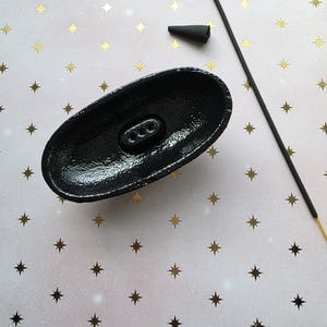 May include: A black, oval-shaped incense holder with three holes, a black incense cone, and a black incense stick with a gold tip. The items are on a pink background with gold star patterns. The holder has a textured surface.