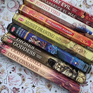 May include: An assortment of HEM incense sticks in various scents, including "Go Away Evil," "Positive Vibes," and "Dragons Blood." The boxes are colorful with text in English and Spanish. The background is a light pattern with crystals, moons, and butterflies.
