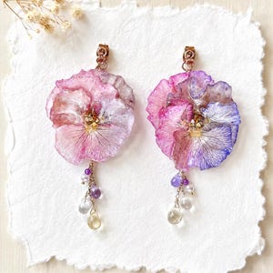 以下が含まれることがあります： ピンクと紫の色合いの押し花パンジーを使用したドロップイヤリングのペア。各花にはゴールドトーンのポストバックが付いており、白いパール、紫のアメジスト、透明なクリスタルを含む小さなぶら下がりビーズのクラスターが添えられています。