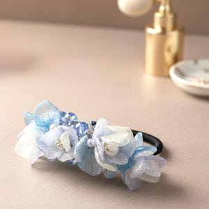 Può includere: Un elastico per capelli con delicati fiori di tessuto blu e bianco a strati e perline trasparenti. La composizione floreale è attaccata a un elastico nero. Accessorio per lo styling dei capelli.