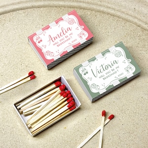 May include: Two matchboxes, one pink and one green, with the text "Amelia WILL YOU BE MY BRIDESMAID?" and "Victoria WILL YOU BE MY BRIDESMAID?" respectively. The matches have red tips. The boxes are on a light-colored surface.