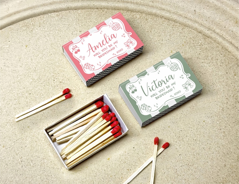 May include: Two matchboxes, one pink and one green, with the text "Amelia WILL YOU BE MY BRIDESMAID?" and "Victoria WILL YOU BE MY BRIDESMAID?" respectively. The matches have red tips. The boxes are on a light-colored surface.