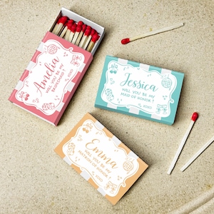 May include: Three decorative matchboxes in pink, teal, and orange, each with a unique name and invitation text. The pink box says "Amelia, will you be my bridesmaid?" The teal box says "Jessica, will you be my maid of honor?" The orange box says "Emma, will you be my matron of honor?"