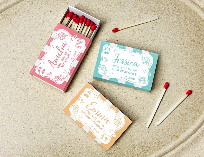 May include: Three decorative matchboxes in pink, teal, and orange, each with a unique name and invitation text. The pink box says "Amelia, will you be my bridesmaid?" The teal box says "Jessica, will you be my maid of honor?" The orange box says "Emma, will you be my matron of honor?"