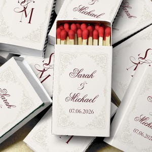 May include: White matchboxes with red-tipped matches inside. The boxes are decorated with floral designs and the names "Sarah & Michael" along with the date "07.06.2026" in burgundy script. The boxes are stacked together.