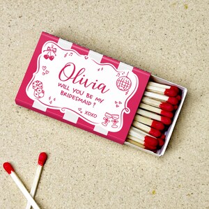 May include: A pink matchbox with white stripes and the text "Olivia, will you be my bridesmaid?" is open, revealing red-tipped matches. Two loose matches with red tips are in the foreground. The box has decorative elements.