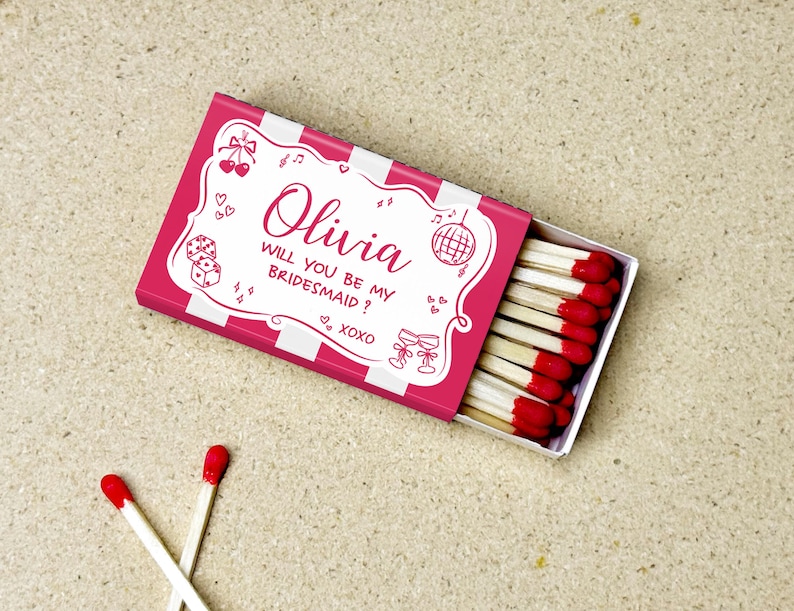 May include: A pink matchbox with white stripes and the text "Olivia, will you be my bridesmaid?" is open, revealing red-tipped matches. Two loose matches with red tips are in the foreground. The box has decorative elements.