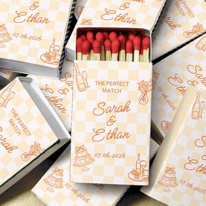 May include: Matchboxes with red-tipped matches, featuring a cream and tan checkered pattern. The boxes are decorated with illustrations of a cake, champagne, and flowers. Text on the boxes reads "The Perfect Match" and names "Sarah & Ethan" with a date.